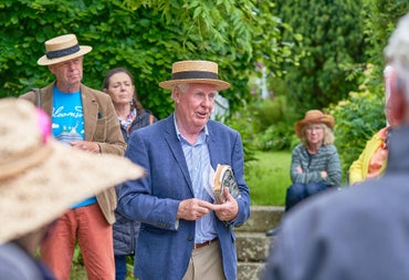 Outdoors a small group of people are around a man giving a talk wearing a straw boater hat.