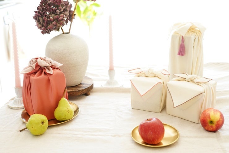 A Group of 4 various sized boxed wrapped in plain cloth with a bow, on a table with plain cloth and some apples and pears.