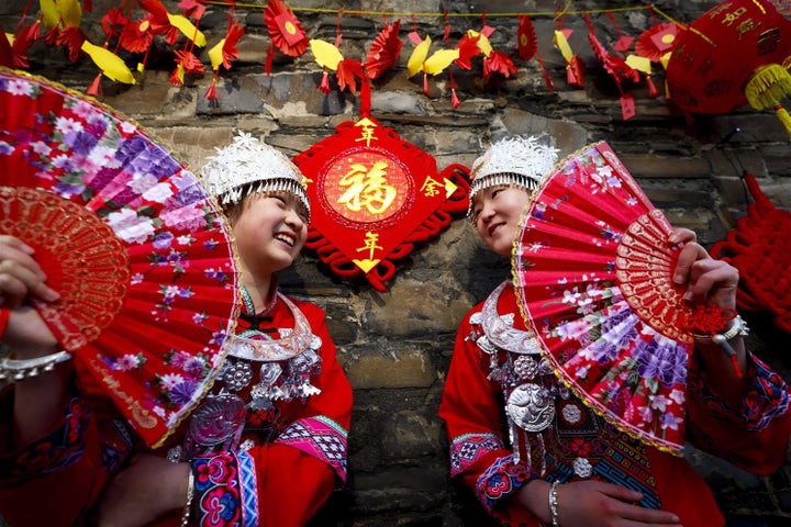 2 people in red national costumes holding large, red fans are smiling at each other, standing in front of a stone wall decorated with red and yellow decorations.
