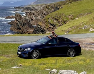 A luxury car and driver parked by a coastal road with rocky coastline in background