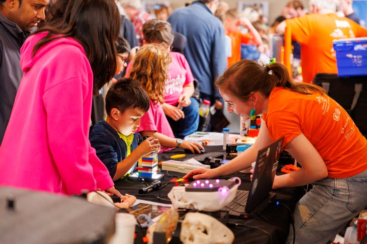 Children and adults gather around a table at a STEM workshop, exploring hands-on activities using lego, microscopes and a laptop.