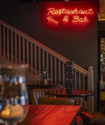 Stairs next to wooden tables and a red neon sign saying bar and restaurant with an arrow pointing upstairs