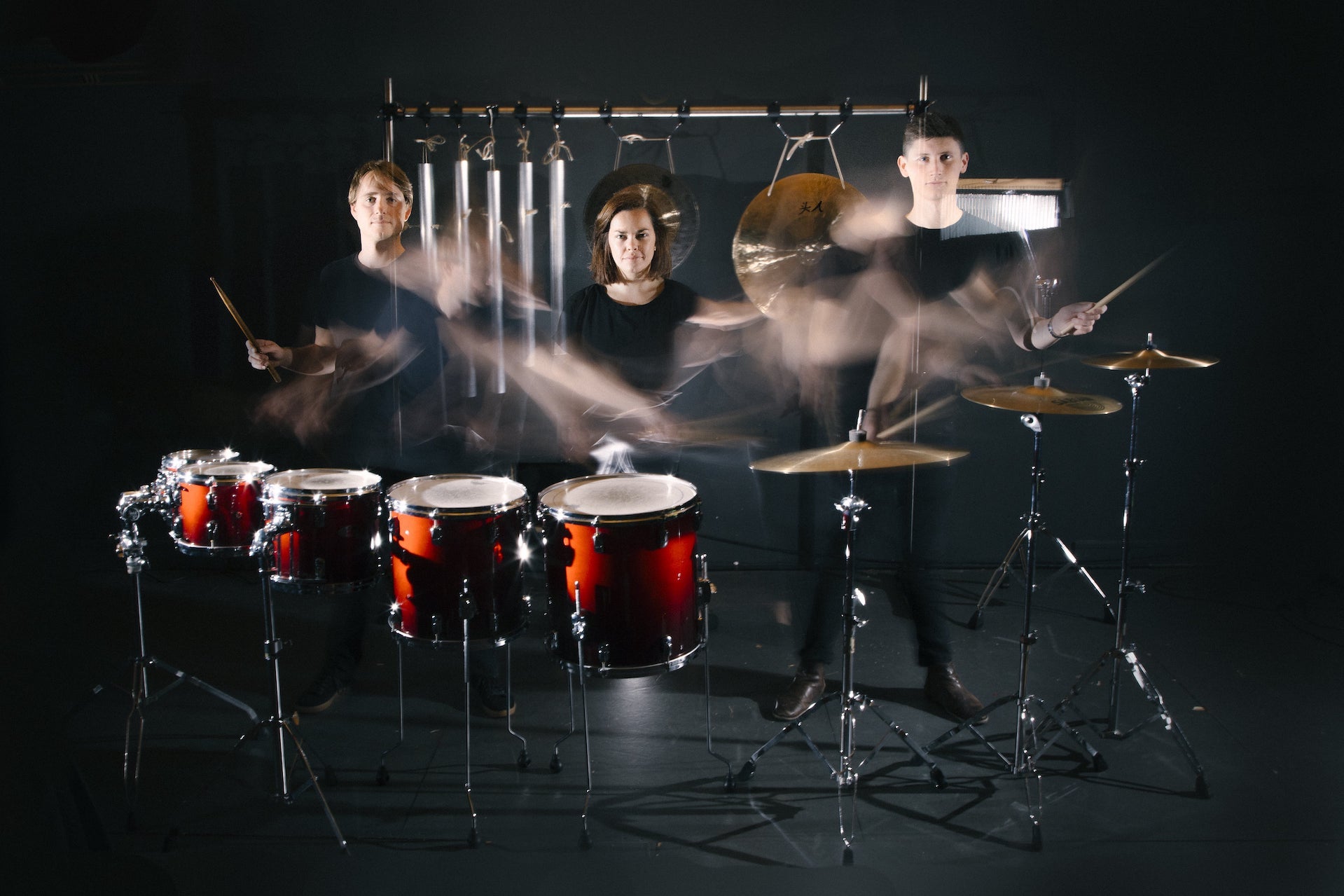 3 people standing behind drums and cymbals on standing with various percussion instruments, their hands blurred with movement