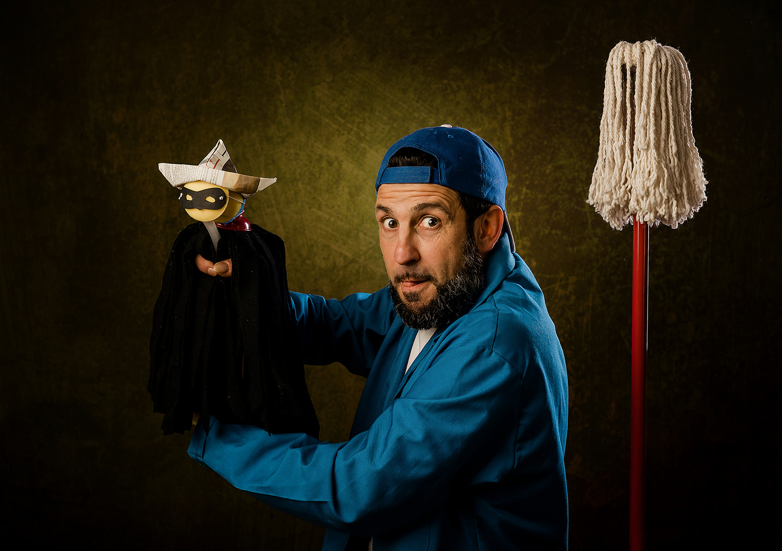 A man in dark blue shirt and cap is holding a small, simple puppet looking surprised, against dark background with an old fashioned mop behind him.
