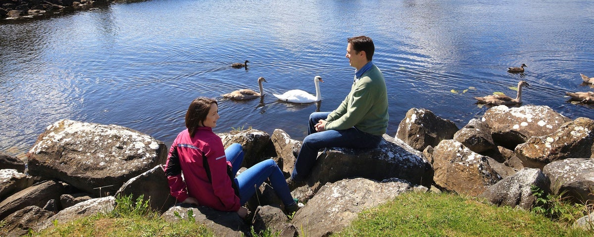 Two people sitting on rocks beside water with swans beside them