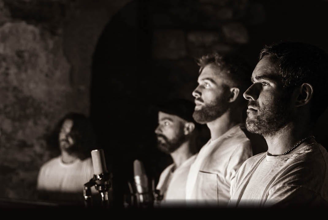 Black and grey shot of 4 men in a line, viewed from the side, fading to blurry in the distance, dark background.