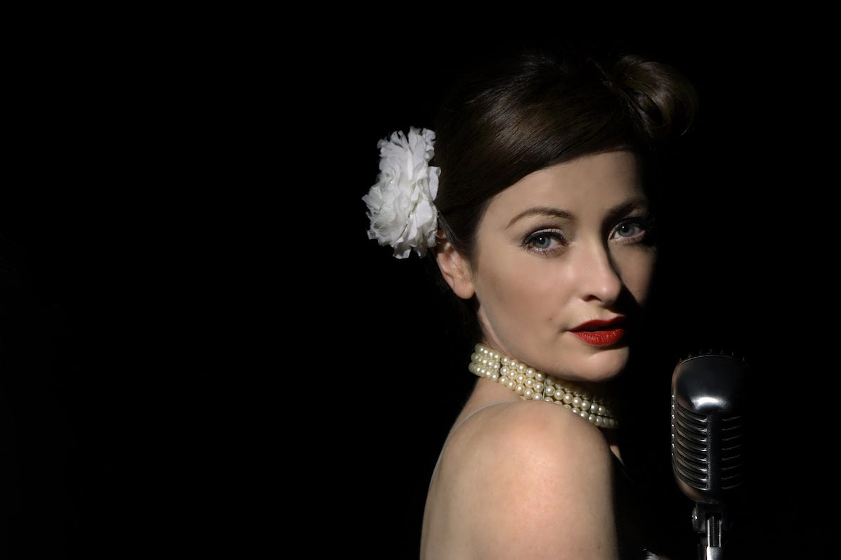 A woman with tied back hair held by white flower is looking over her bare right shoulder at camera with an old style mic in front of her, against black background.