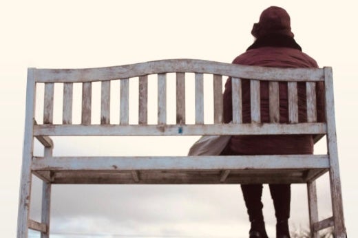 'An tÉalú' Ceoldráma Nua le Seán Óg Ó Duinnín - rear view of person in winter coat and hat seated at one end of a wooden bench.