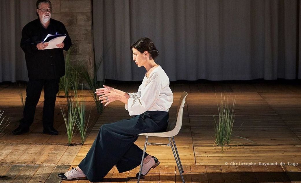 An image from Sopro. Two people on stage, one is sitting the foreground another is standing in the background with a script in hand.