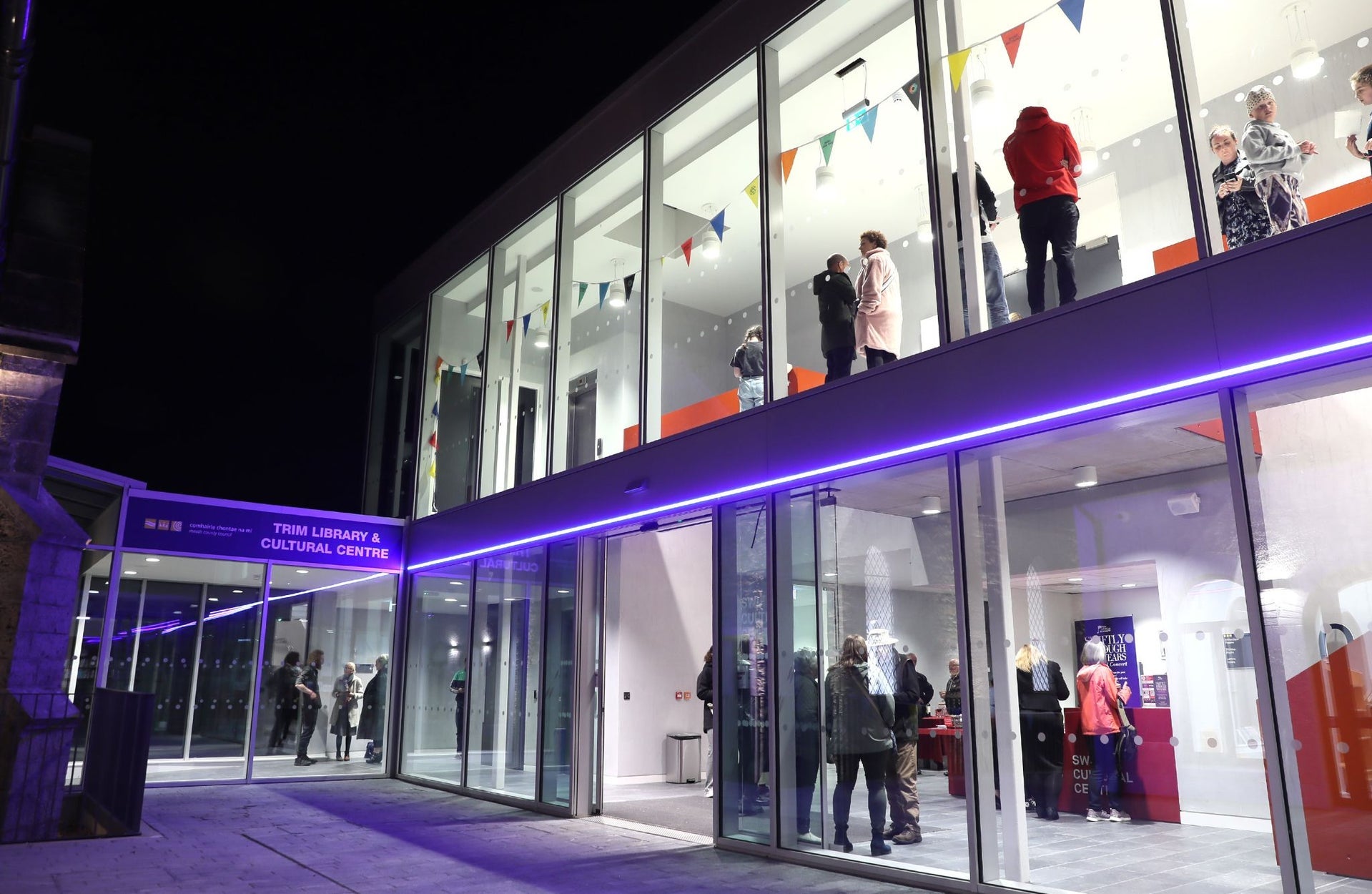 The exterior of the Swift Cultural Centre at night with people inside a well lit lobby and waiting area
