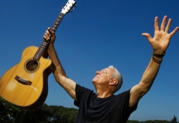 A man with arms outstretched, holding up a guitar in his right hand against blue sky