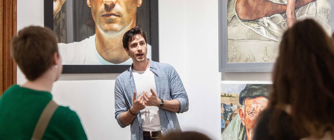 A man is talking using is hands, standing in front of wall of art picture.