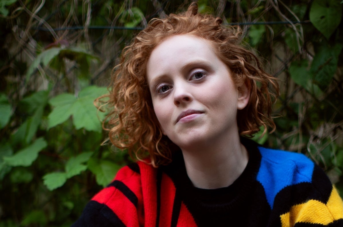 Jessie Grimes, presenter, gently smiling woman in colourful jumper against background of green plants