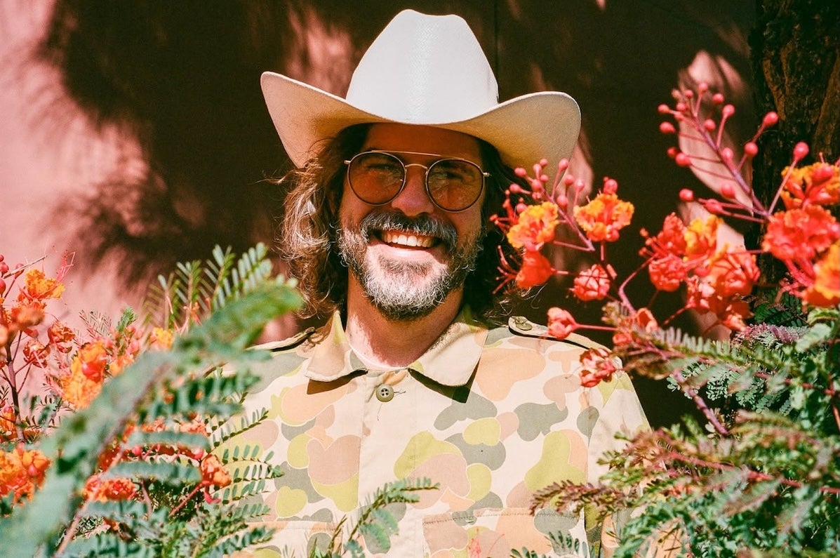A wide smiling man in a cowboy hat, partially obscured by branches and foliage.