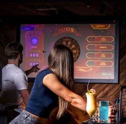 Person leaning on a counter with drinks as another person points at a digital dart board