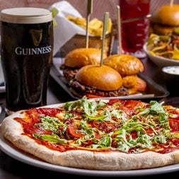 Pint of stout on a table next to a pizza sliders and a basket of chips in the background