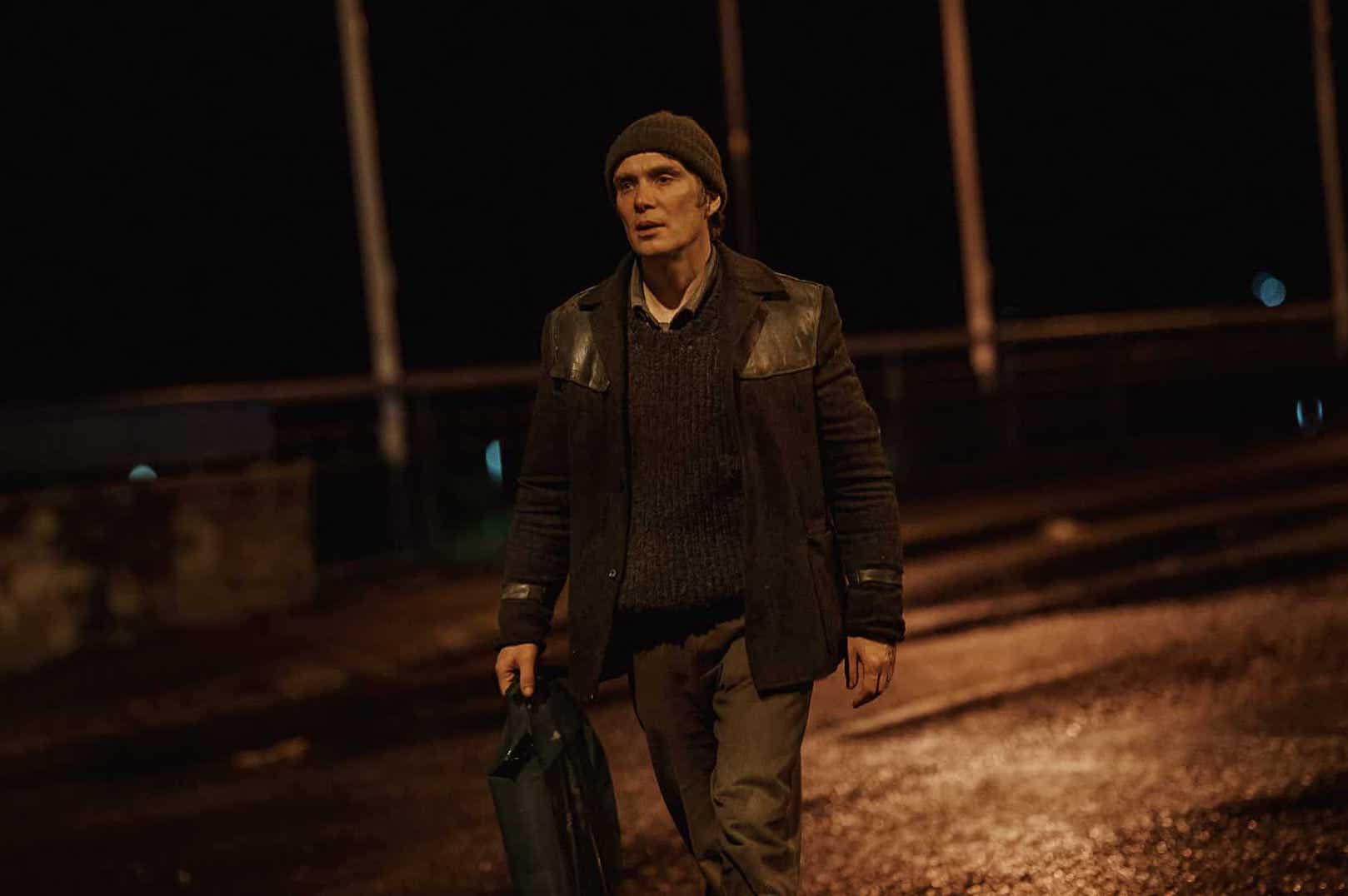 A man in well worn clothes is walking along a road at night.