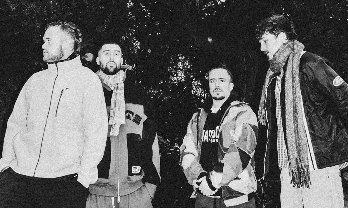 Slightly grainy black and white photo of 4 men with outdoor jackets on and trees in the background.