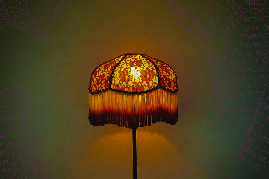 A foral lampshade with long, fringed edge, lit against dull green background.