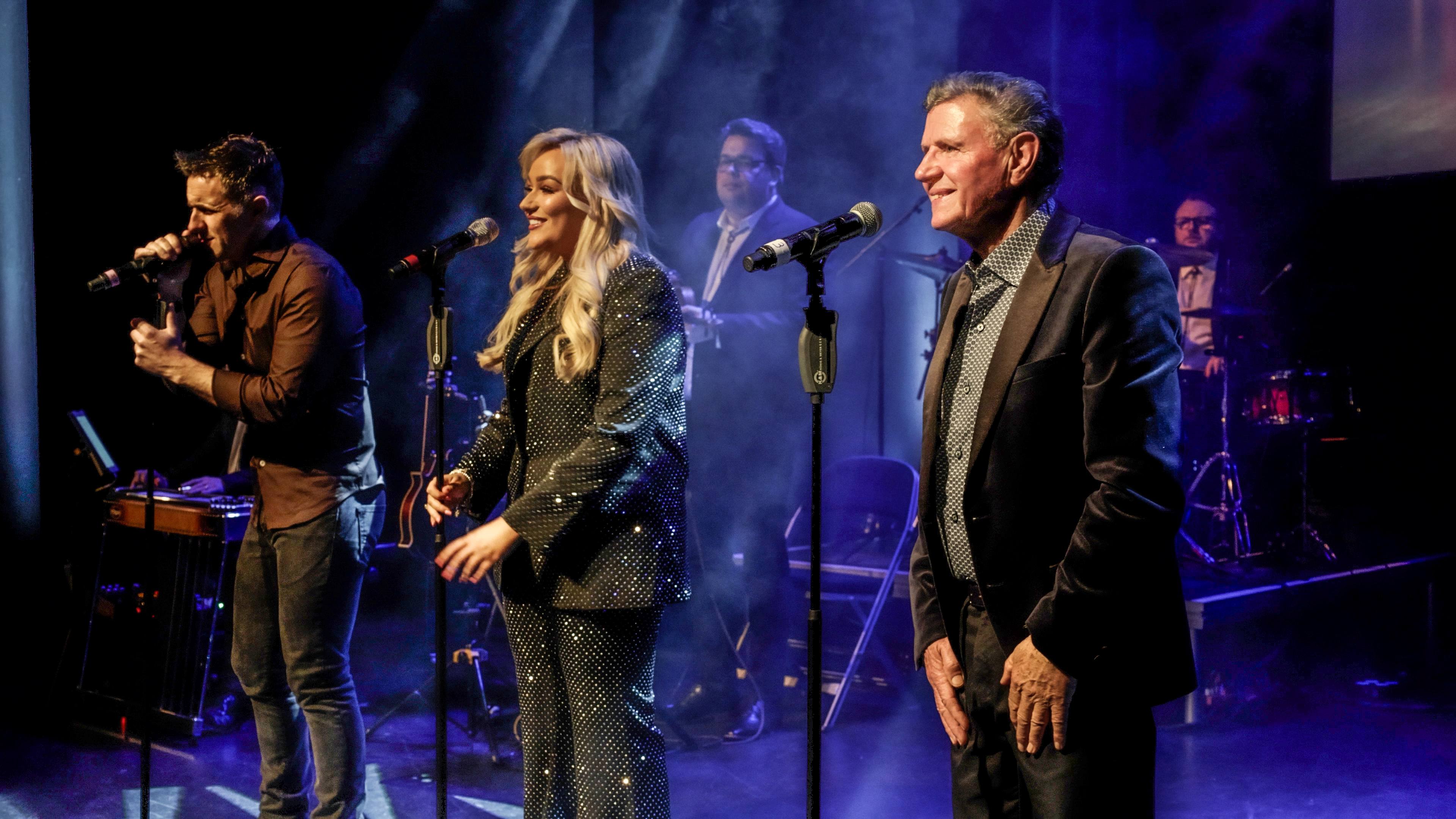3 people standing with mics on a stage singing with band behind.