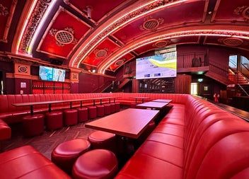 A theatre style lounge with luxurious red leather booth seating and ornate decorative ceilings with large screens throughout
