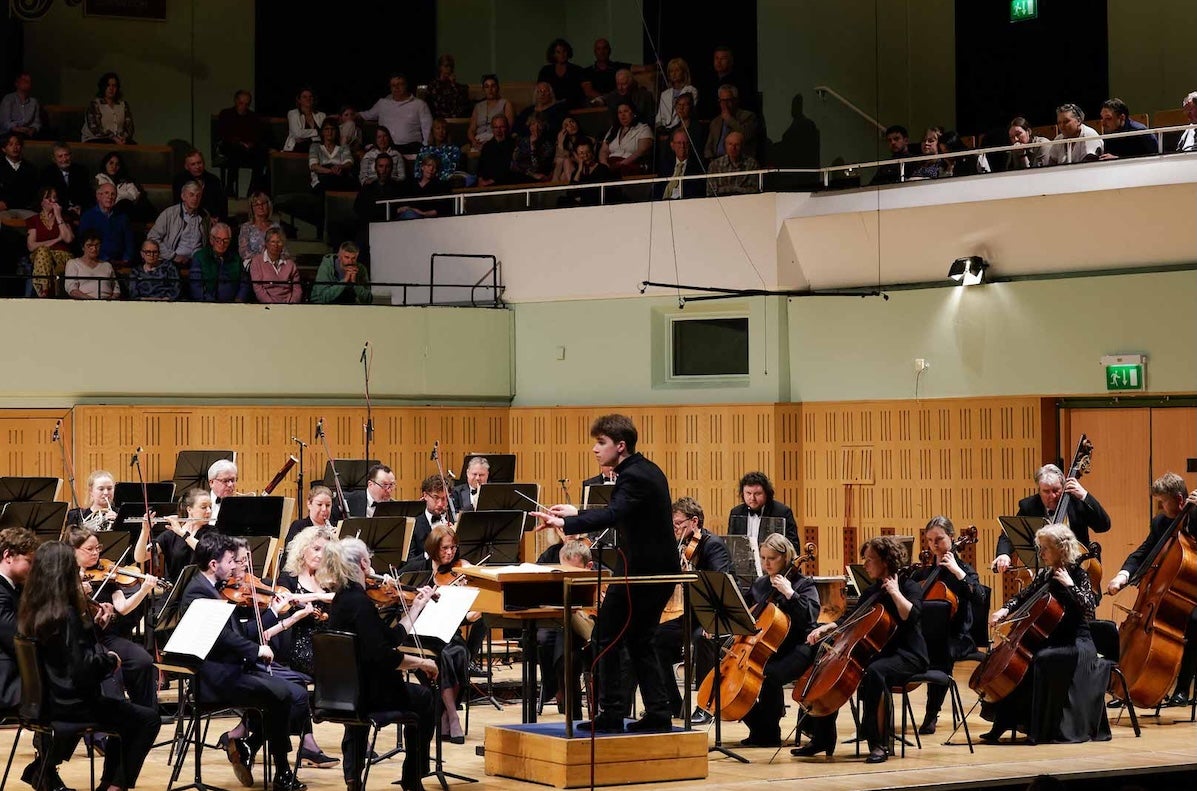 Part of an orchestra playing and being conducted by man in black with part of audience visible seated above.