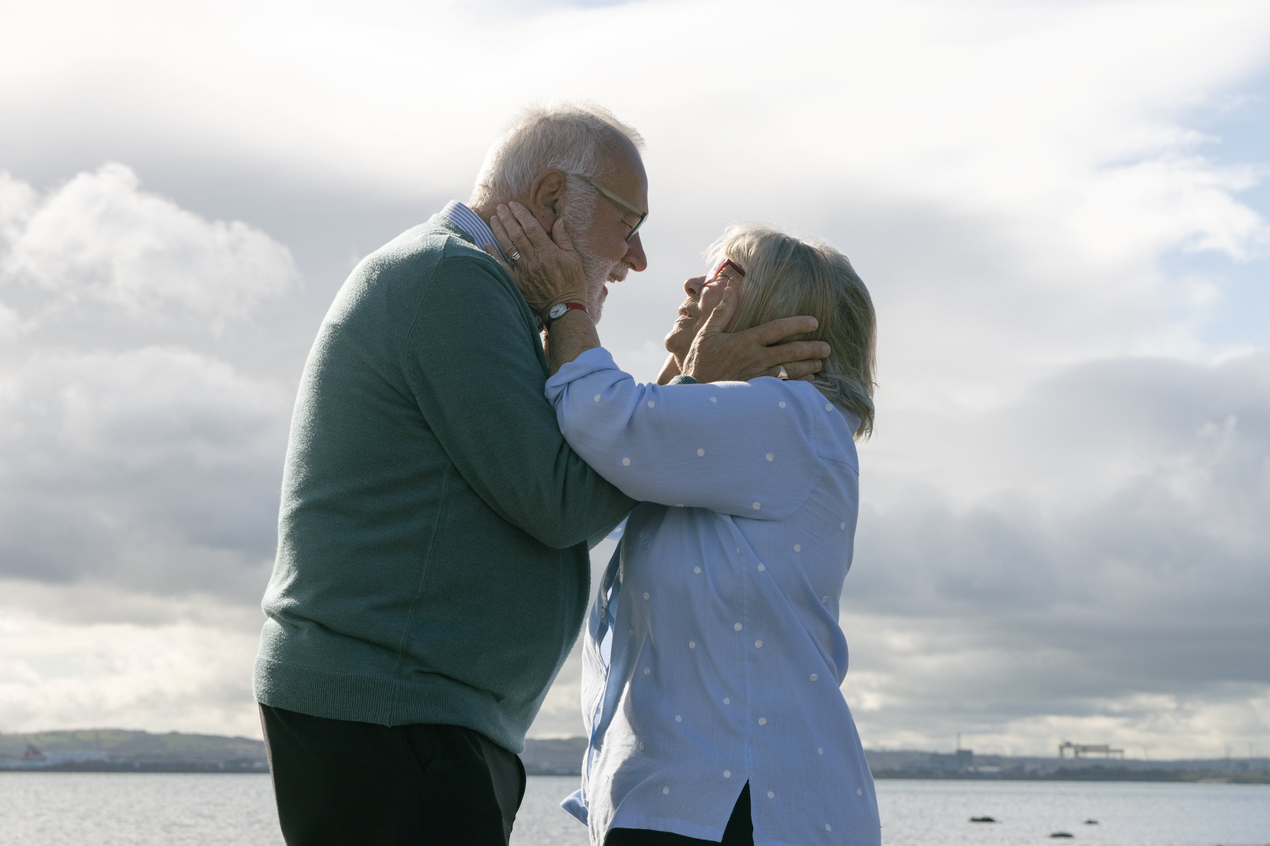 An old man and woman are facing each other, holding each other's heads tenderly.