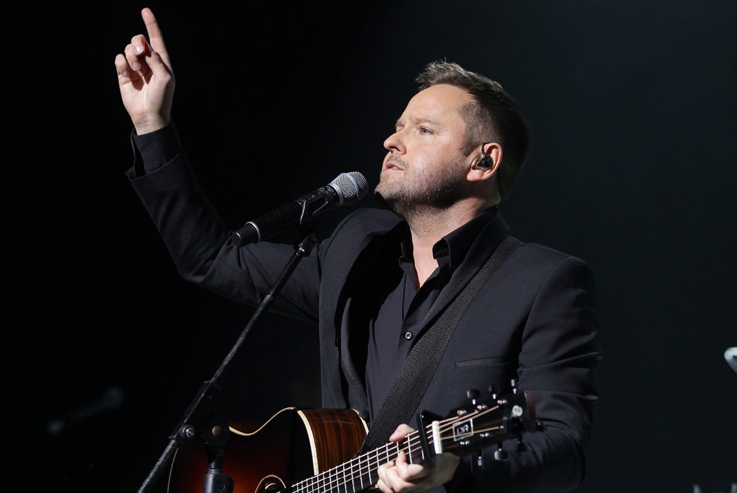 A man playing guitar with a mic on a stand and his right arm raised pointing up into the air.