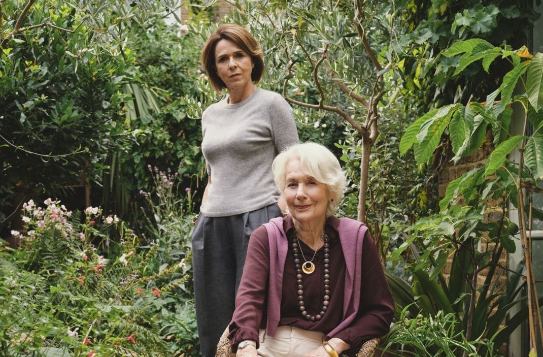 Meredith sits in a rattan chair in the garden, while her daughter Noelle stands behind her.