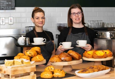 Two people standing behind a counter with pastries on display holding cups and a tray
