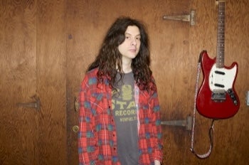 A person with long dark hair and checked shirt, standing beside a guitar hanging on the wall.