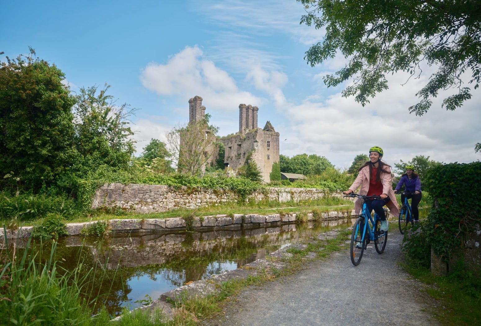 Two cyclists on a path by a river with the ruins of a castle in the background