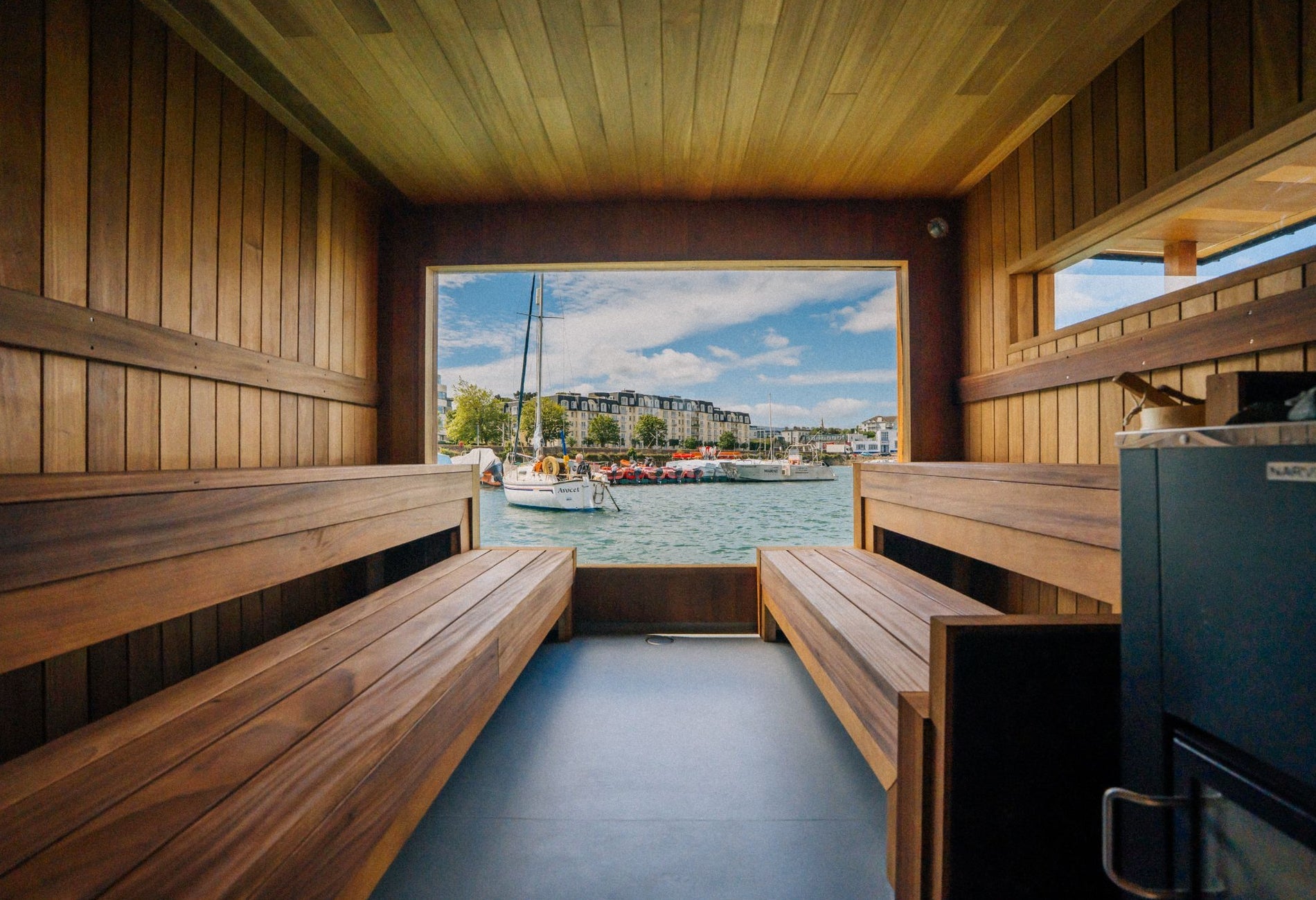 Wood lined sauna interior with bench seating overlooking a harbour through a large open window