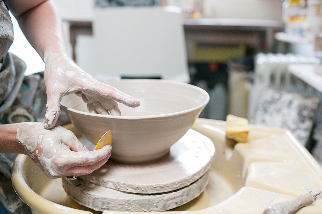 Clay covered hands working on a clay bowl on a potter's wheel.