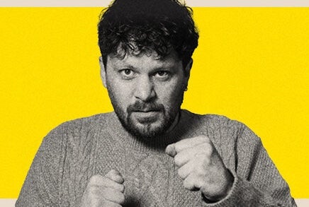 A man looking a bit fierce holding up his hands in fists in front of him, against bright yellow background.