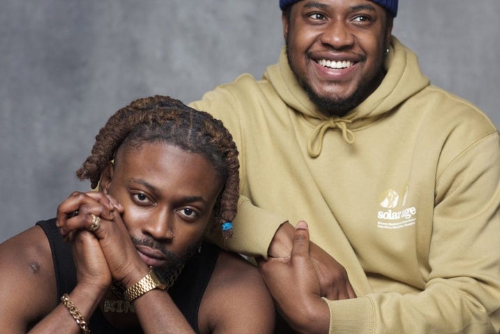 Musical Strings A Portrait Exhibition by Emily Quinn. 2 men, one resting on the shoulder of the other, smiling broadly against plain grey background.