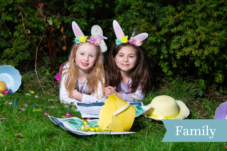 2 young children wearing white rabbits ears are laid down on grass with easter bucket and eggs in front of them and a yellow hat.
