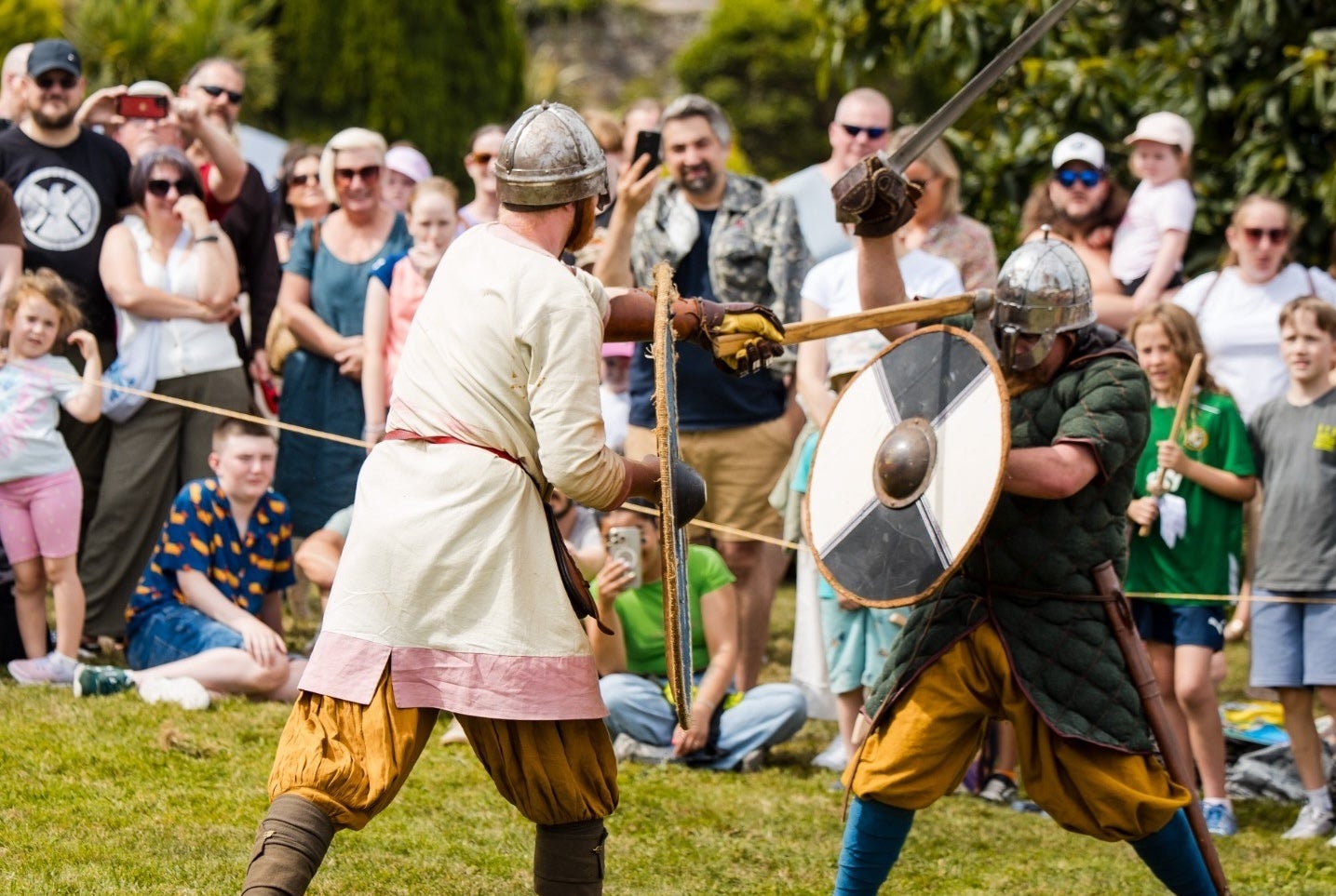 Viking Battle Re-enactment, 2 men in viking style clothes with metal helmets fighting with round shields and wooden swords with crowd watching on.