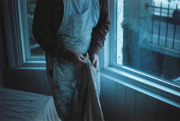 Person in old apron holding a item made of cloth, standing by a window, their head not visible.