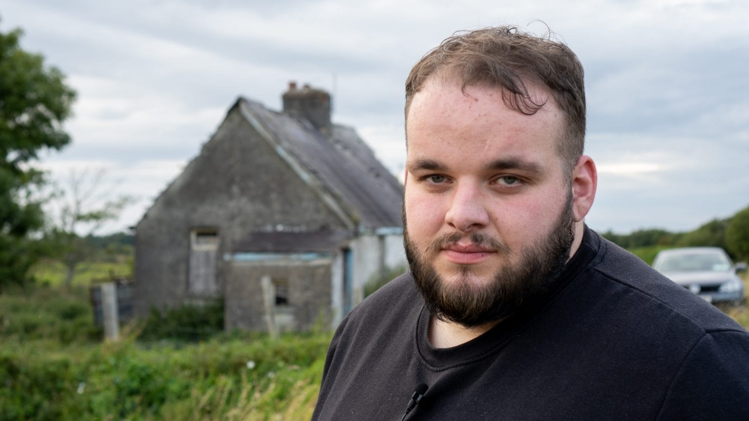 Writing Home is a documentary film by Arcade. A man with a beard wearing a black top looking serious with an old, small stone building in the background in the countryside.