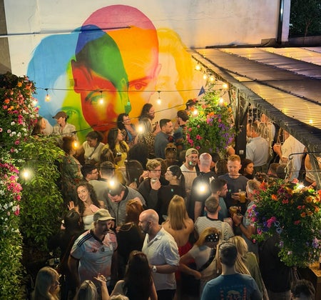 Crowds gathered in the outdoor space of a bar beneath string lights and a colourful mural backdrop