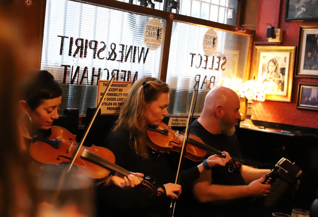 Three musicians sitting in a pub playing instruments with a window behind them