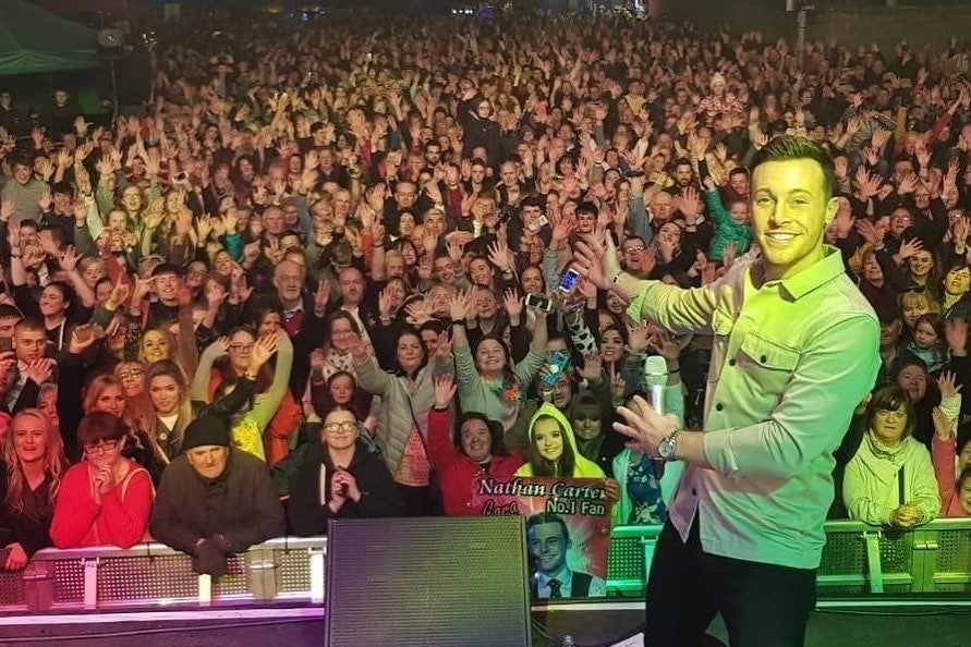 A view from the back of a stage looking out over a large crowd at night with a man to one side smiling, pointing at the audience.