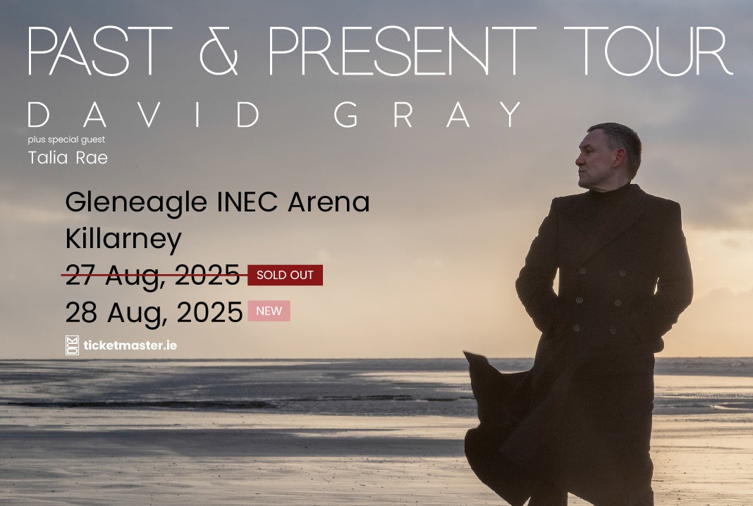 A man in dark long coat at dusk standing at the edge of the sea on a beach.