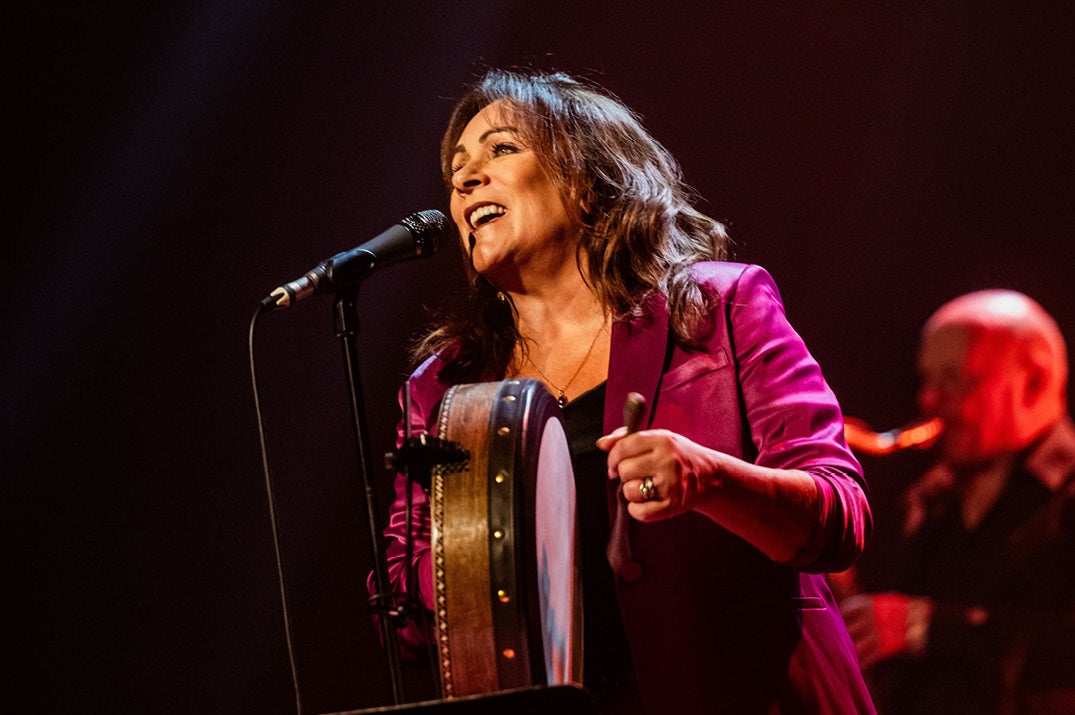 A woman singing into a mic on a stand holding a small round drum with blurred musician in the dark background.