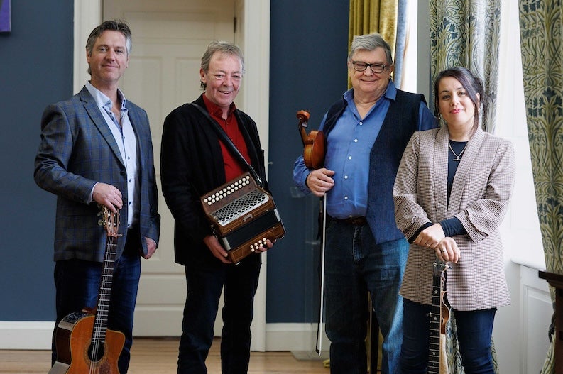 4 smiling musicians standing with instruments beside a window with curtains