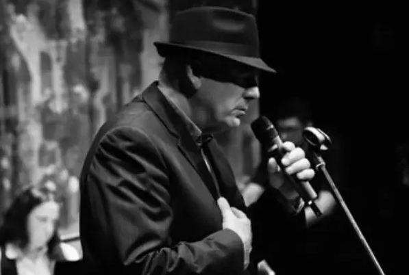 Black and white photo of man in dark suit and hat, viewed from the side, holding a mic on a stand, eyes in shadow from brim of hat.