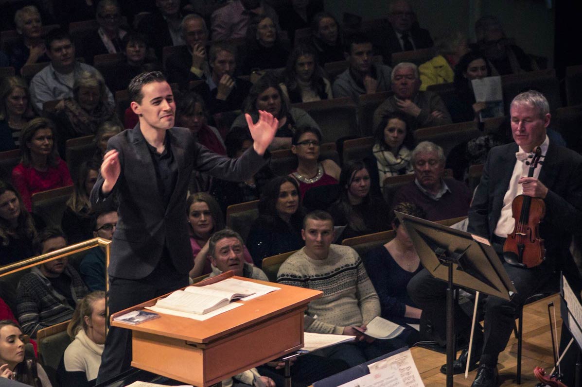 A man dressed in black is conducting with audience behind him and a man in evening dress seated holding a violin visible to the right.