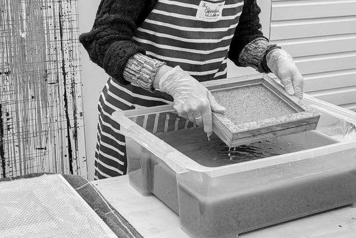 A pair of hands lifting a small wooden square frame over a large plastic container holding a dark liquid.