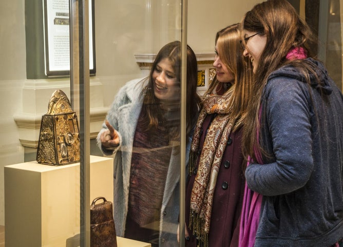 3 women are smiling, one pointing at a gold object in a glass display cabinet which they are gathered around.